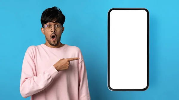 ピンクのトレーナーと眼鏡でショックを受けた後ろ男で大きなスマートフォンで彼の隣に白い空の画面 青のスタジオの背景 モックアップ コピースペース お勧めします素晴らしいモバイルアプリ — ストック写真