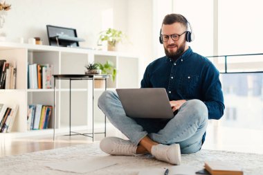 Erkek Freelancer İnternet 'i tarıyor Laptop kullanıyor, kulaklık takıyor ve evde oturuyor.