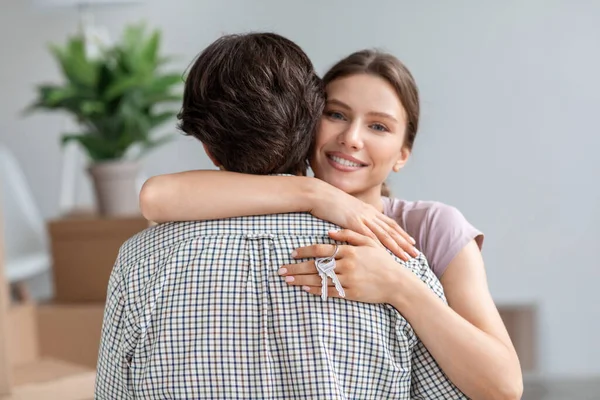 Alegre millennial europeo hembra en casual abrazo marido muestra las llaves en la habitación con embalado cajas de cartón —  Fotos de Stock