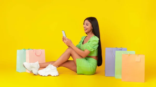 Gelukkige vrouw met smartphone, zittend op de vloer met boodschappentassen — Stockfoto