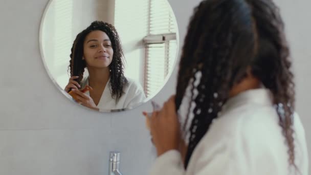 Peinado doméstico. Joven dama americana rizada y pacífica en albornoz peinando el cabello, mirando al espejo en el baño — Vídeo de stock