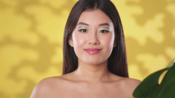 Naturlig skönhetsvård. Porträtt av vacker asiatisk kvinna med kreativ makeup poserar till kameran med gröna monstera blad — Stockvideo