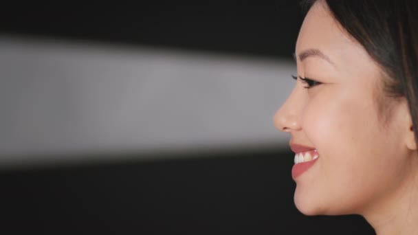 Профиль портрет позитивной азиатской женщины, смотрящей в сторону на пустое пространство, черный фон с лучом света, замедленная съемка — стоковое видео
