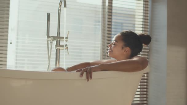 Vattenspa hemma. Ung afrikansk amerikansk kvinna avkopplande i varmt bad, liggande i badkar med hällande vatten på morgonen — Stockvideo