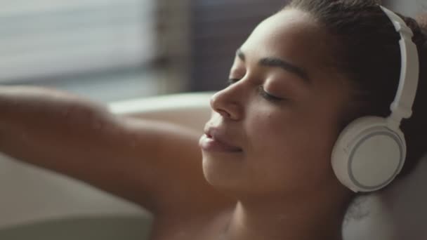 Menutup diri dari wanita muda Afrika-Amerika yang mendengarkan musik melalui headset nirkabel, bersantai di kamar mandi panas dengan mata tertutup — Stok Video