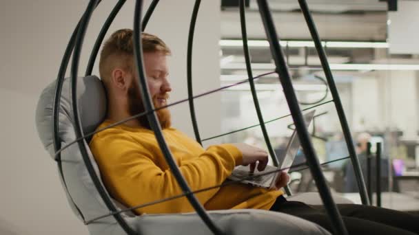 Hombre usando el ordenador portátil de trabajo en línea sentado en la silla oscilante interior — Vídeos de Stock