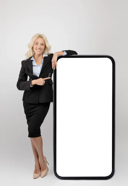 Jovem mulher de negócios feliz apoiando-se no smartphone gigante com tela branca vazia e apontando para o grande dispositivo, mockup — Fotografia de Stock