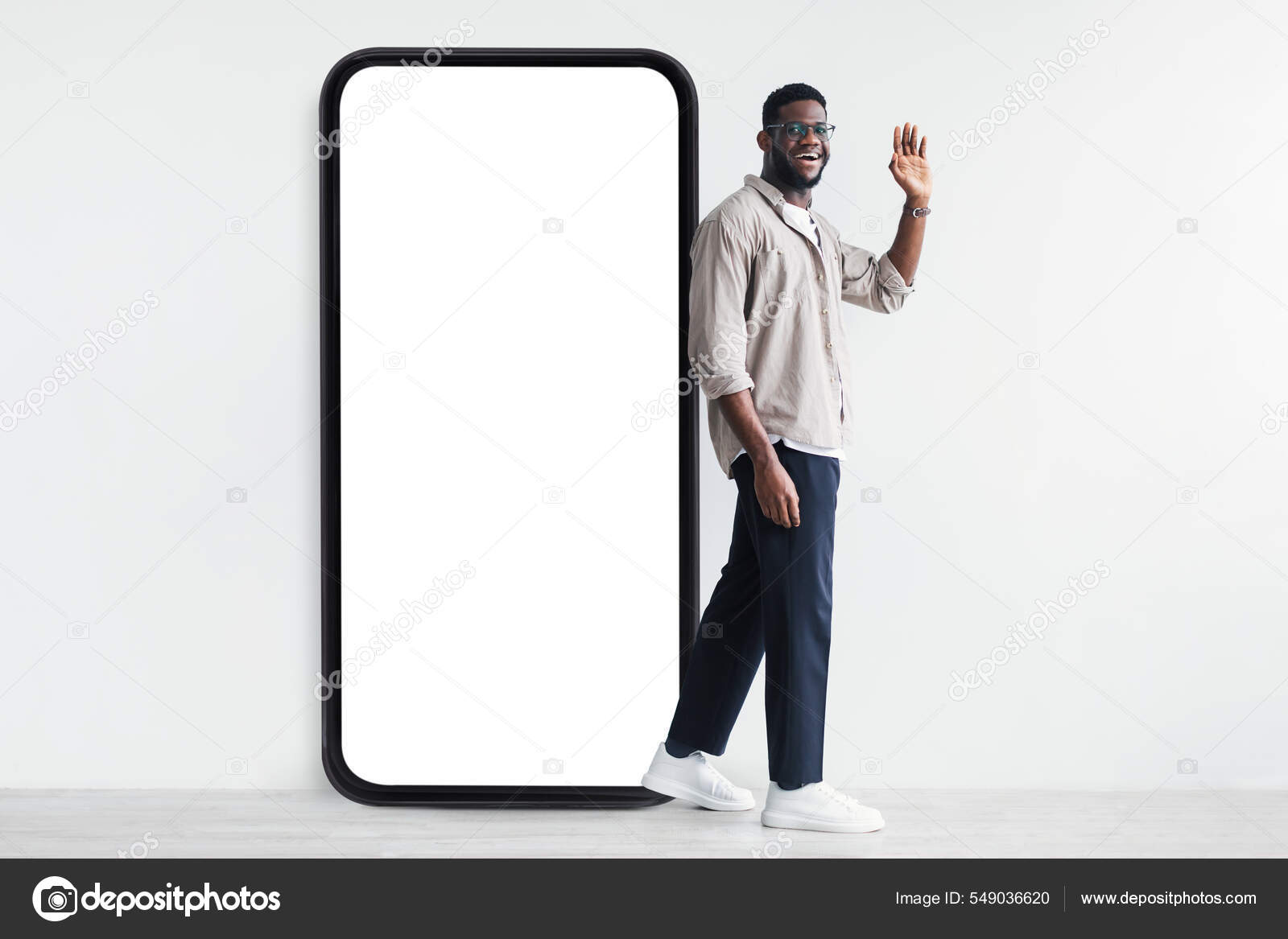 Счастливый черный парень стоит рядом с гигантским смартфоном с белым экраном и машет привет в камеру, макет стоковое фото ©Milkos 549036620