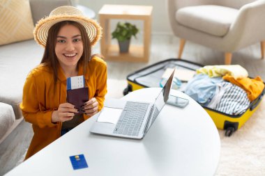 Pasaportu ve bileti olan neşeli genç bir bayan bilgisayarını kullanarak evinden online seyahat acentesinde tatil satın alıyor.
