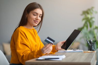 Tablet bilgisayarlı ve kredi kartlı neşeli genç bayan evde uzaktan banka hizmeti kullanarak online sipariş veriyor.