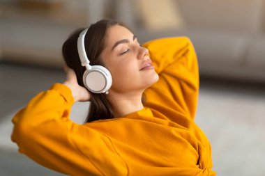Kablosuz kulaklıkla müzik dinleyen, elleri başının arkasında dinlenen, sakin, beyaz bir kadın.