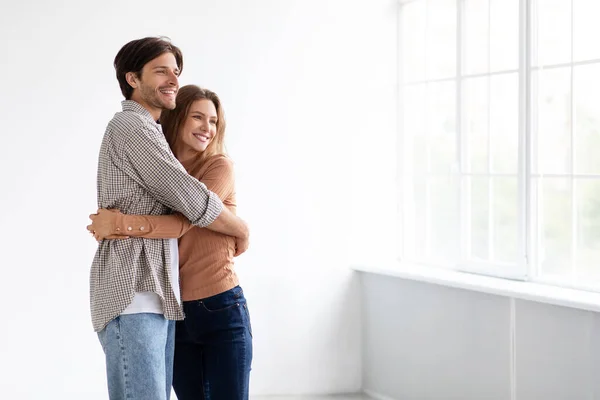 Alegre abrazo caucásico milenario masculino y femenino, mirando a la ventana en la habitación vacía del nuevo apartamento, resplandor solar —  Fotos de Stock