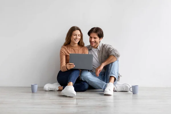 Feliz sonriente joven europeo hombre y la señora miran portátil planificación de nuevo interior, sentado en el suelo en la habitación vacía — Foto de Stock