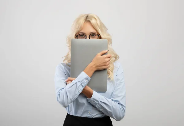 Удивительная молодая деловая женщина покрывает лицо ноутбуком, носит формальную одежду и позирует на светлом фоне студии — стоковое фото
