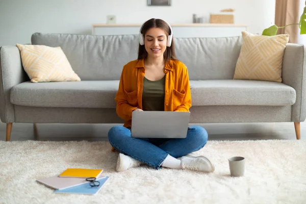 Mujer joven positiva en los auriculares que trabajan en el ordenador portátil, sentado con las piernas cruzadas en el suelo en casa, longitud completa — Foto de Stock
