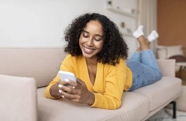 Millennial senhora negra usando celular, deitado no sofá em casa, trabalhando ou estudando remotamente — Fotografia de Stock