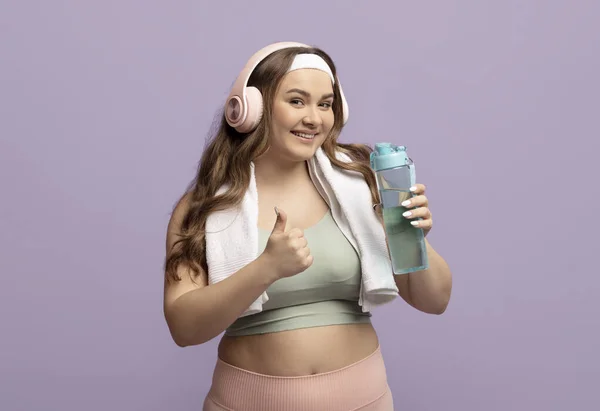 Happy plus size european millennial lady in sportswear with towel in headphones with bottle of water