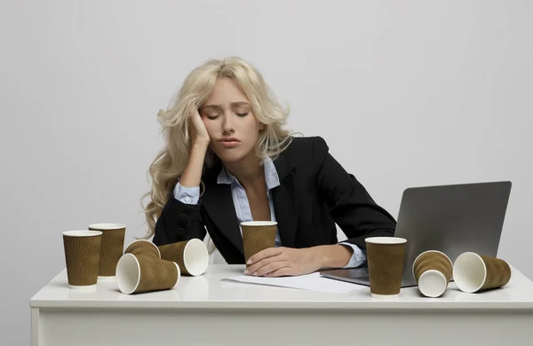 Trabajadora de oficina agotada rodeada de tazas de café vacías durmiendo en el lugar de trabajo sobre fondo de estudio gris claro — Foto de Stock