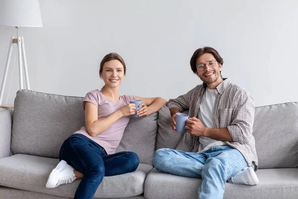 Feliz millennial chico caucásico en gafas y la mujer en el descanso casual en el sofá, beber café —  Fotos de Stock