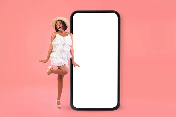 Playful black woman jumping near big cellphone with empty screen for mobile offer or application ad over pink background — Stock Photo, Image