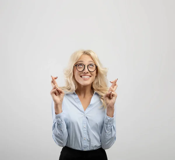 Emocionada mujer de negocios rubia en ropa formal cruzando los dedos para la buena suerte y mirando hacia arriba sobre fondo gris claro, cosecha —  Fotos de Stock