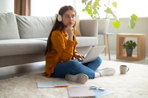 Videollamada en línea. Mujer joven reflexiva en auriculares sentados en el suelo, utilizando el ordenador portátil, teniendo conferencia virtual en interiores — Foto de Stock