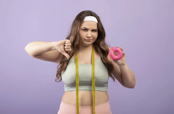Displeased upset plus size european young female in sportswear with measuring tape show donut and thumb down — Stock Photo, Image