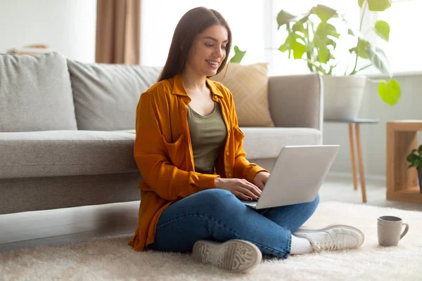 Πλήρες μήκος του ευτυχισμένη νεαρή γυναίκα κάθεται σταυρωτό πόδι στο πάτωμα με φορητό υπολογιστή, εργασίας ή μελέτης σε απευθείας σύνδεση στο σπίτι — Φωτογραφία Αρχείου