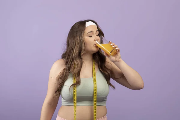 Lugn söt plus storlek ung kvinna i sportkläder med slutna ögon och mäta tejp dricka glas apelsinjuice — Stockfoto