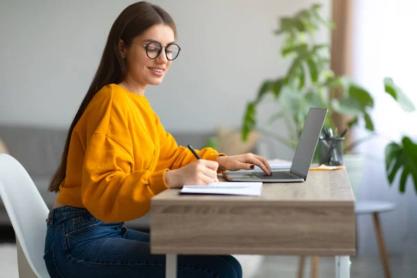 Glimlachende jonge vrouw in bril aan het bureau, laptop aan het werk, schrijfblok thuis, vrije ruimte — Stockfoto