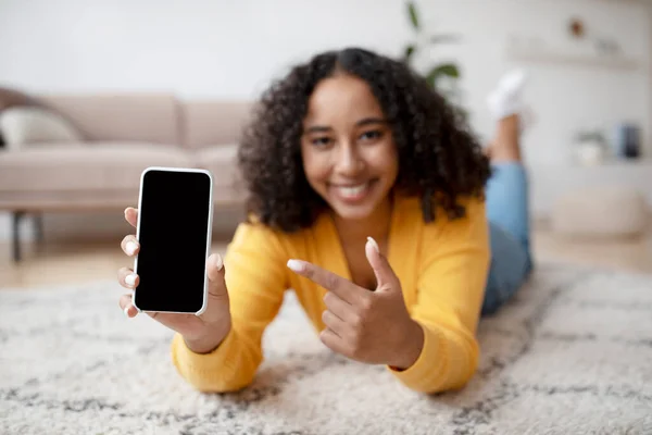 Giovane donna afroamericana felice che punta lo smartphone con schermo vuoto, sdraiata sul pavimento a casa, mockup — Foto Stock