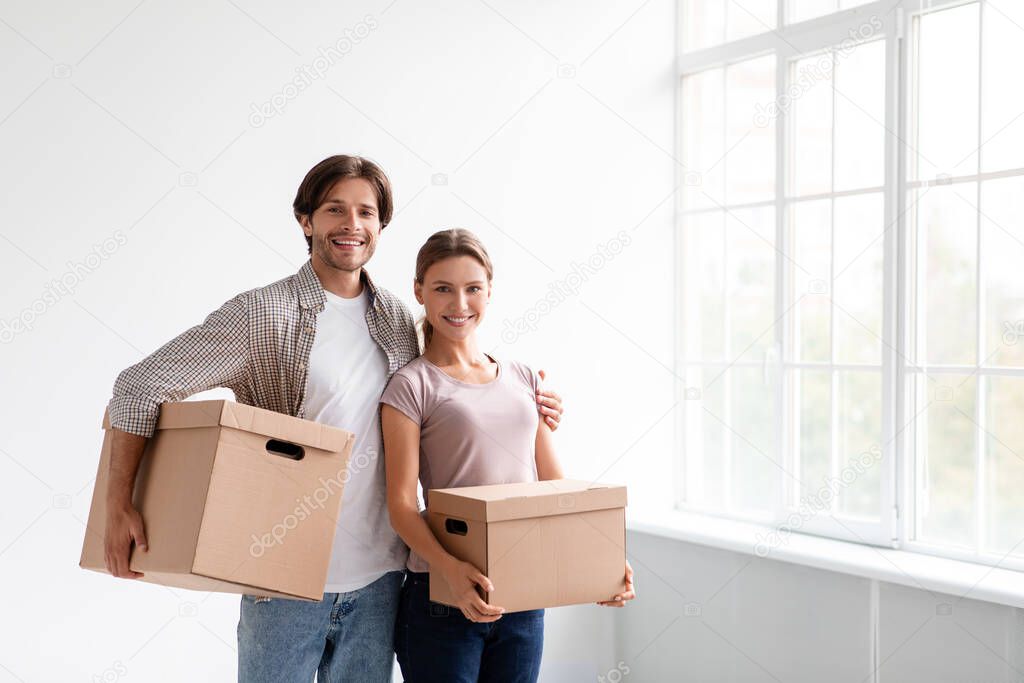 Glad millennial caucasian man and woman in casual carry boxes, hugs and enjoy renting new flat in empty room