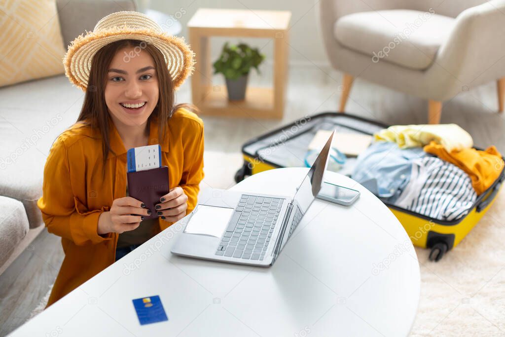Cheerful young lady with passport and ticket using laptop to buy tourist vacation at online travel agency from home