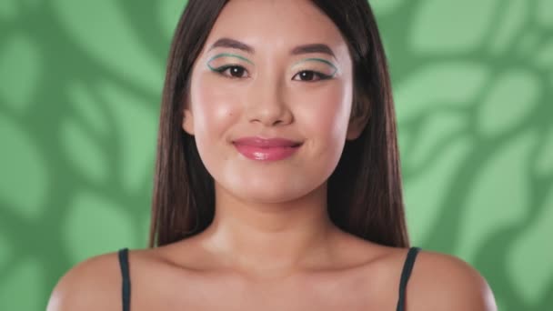 Flirt. Junge verspielte asiatische Frau mit kreativem Make-up schickt Pusteblume in die Kamera und flirtet über grünen Hintergrund — Stockvideo
