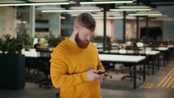 Vousatý muž používající smartphone textové nosící sluchátka stojící na pracovišti
