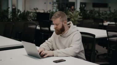 Sakallı adam iş yerinde dizüstü bilgisayar kullanıyor. İçerideki masada oturuyor.