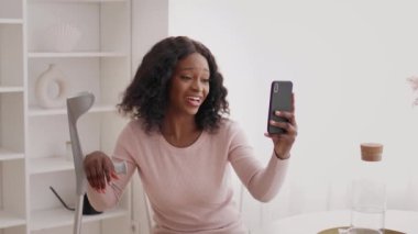 Koltuk değnekli siyah kadın içeride akıllı telefon kullanarak video görüşmesi yapıyor.