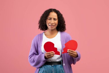 Mutsuz genç siyahi kadın, elinde iki parça kesilmiş kağıt kalp ile pembe stüdyo arka planında ağlıyor.