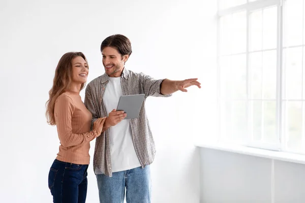 Heureux homme et femme européenne millénaire heureux planification nouvel intérieur avec tablette dans la chambre vide de nouvel appartement avec fenêtre — Photo