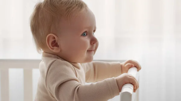 Porträtt av bedårande liten bebis stående i spjälsängen och leende åt sidan, tittar med nyfikenhet, panorama, tomt utrymme — Stockfoto