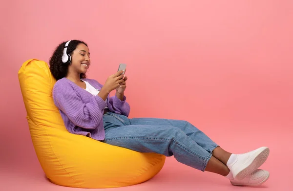 Femme noire avec écouteurs et smartphone écoutant de la musique, appréciant le livre audio, assise dans une chaise de sac de haricot — Photo