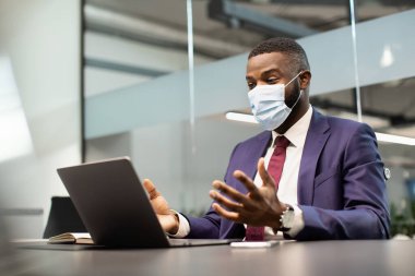 Yüz maskeli siyahi işadamı dizüstü bilgisayarda çalışıyor, konferans yapıyor.