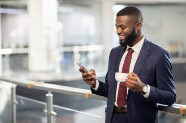 Neşeli siyah yönetici kahve içiyor, cep telefonu kullanıyor, kopyalama alanı kullanıyor.
