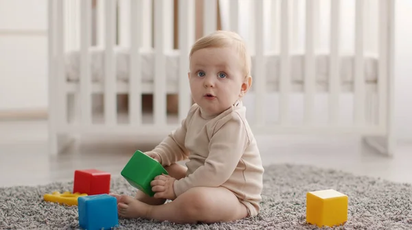 Malé zvědavé miminko si hraje s barevnými kostkami, sedí na podlaze v ložnici, se zájmem hledí nahoru, panorama — Stock fotografie