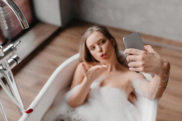 Jonge vrouw rusten in bad en het nemen van selfie op mobiele telefoon, liggend in de badkamer en blazen lucht kus naar webcamera — Stockfoto