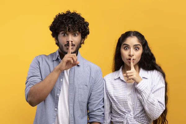 No digas ni una palabra. Hombre y mujer indios manteniendo los dedos en los labios, haciendo señal de silencio, mirando a la cámara, fondo amarillo — Foto de Stock