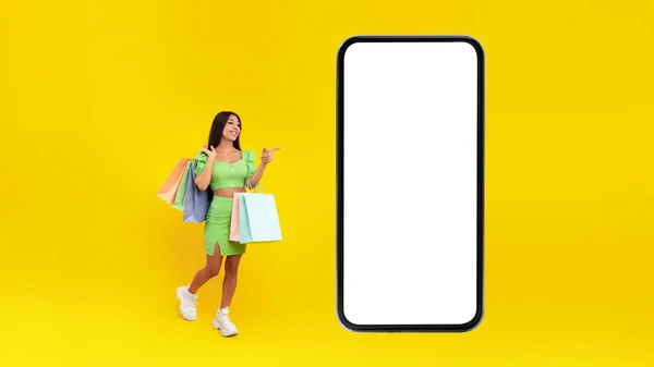 Женщина указывает на белый пустой экран смартфона держа сумки — стоковое фото