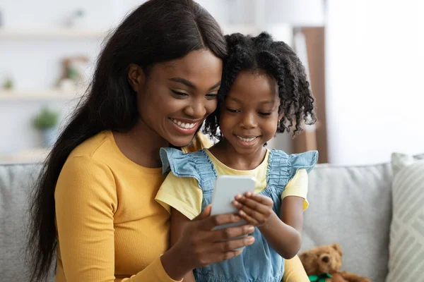 Счастливая афроамериканская мама и маленькая дочь отдыхают со смартфоном дома — стоковое фото