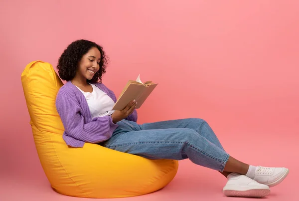 Relaxed νεαρή μαύρη γυναίκα κάθεται στην καρέκλα τσάντα φασόλι με ανοιχτό βιβλίο πάνω από ροζ φόντο στούντιο, κενό χώρο — Φωτογραφία Αρχείου