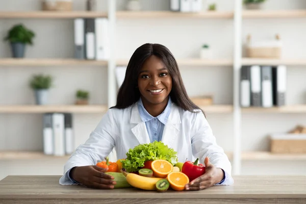 Портрет счастливого африканского диетолога в халате, сидящего со свежими фруктами и овощами на столе — стоковое фото